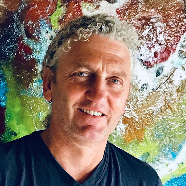 Portrait of an individual standing in front of a vibrant abstract painting with rich textures and dynamic color transitions, reflecting the expressive action‑painting style associated with the artistic work of Jürgen Bauert.